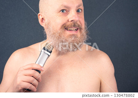 Man grooming his beard with an electric trimmer against a dark background 130478802