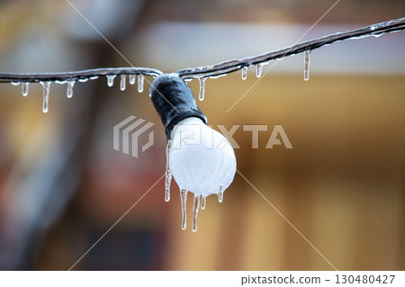 Icicles drip from a light bulb on a frosty wire, set against a warm, blurred backdrop Icicles drip from a light bulb on a frosty wire, set against a warm, blurred backdrop 130480427