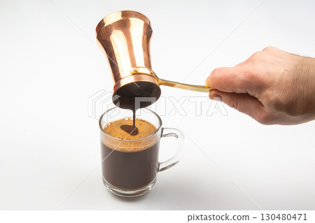 A hand pours Turkish coffee from a traditional copper cezve into a clear glass cup 130480471