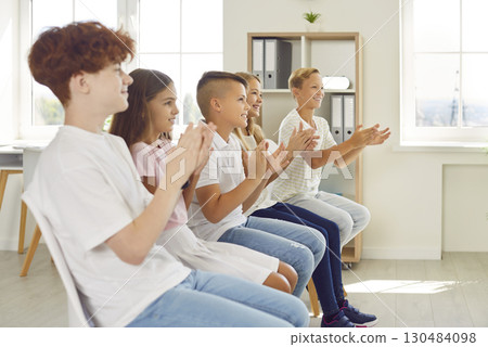 Happy children sitting in a row on chairs and clapping hands at a school event in classroom Happy children sitting in a row on chairs and clapping hands at a school event in classroom 130484098