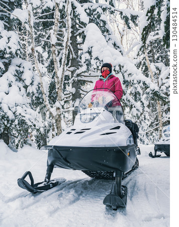Young woman dressed in warm winter clothing navigates a snowmobile along a snowy trail surrounded by tall trees. 130484514