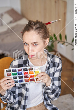 Caucasian young adult female playfully holding watercolor set with paintbrush 130486994