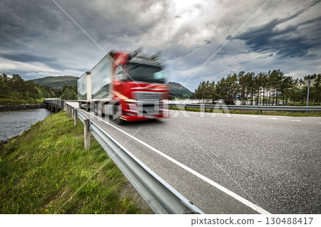 Red semi truck is driving down a road with a cloudy sky in the background. Red semi truck is driving down a road with a cloudy sky in the background. 130488417
