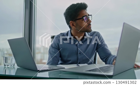 Young business executive sitting at desk with two laptops, documents and glass of water, looking away and thinking about future projects in modern office with big window 130491302