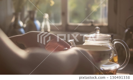 Woman serving blooming tea in transparent teapot, sunlight illuminating warm interior setting near window, creating peaceful afternoon mood 130491338