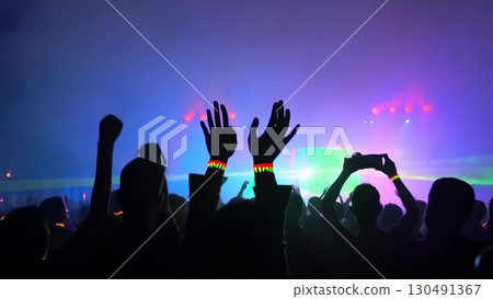 Silhouetted concert crowd raising hands with illuminated wristbands, recording energetic live music performance against bright stage lighting 130491367