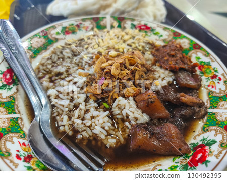Traditional Indonesian rawon beef soup with rice, served in a floral-patterned bowl, topped with fried shallots and sambal, accompanied by crispy crackers. 130492395