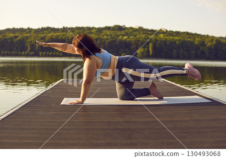 Overweight woman is doing plank exercise while practicing yoga on lake in nature Overweight woman is doing plank exercise while practicing yoga on lake in nature 130493068