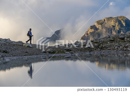 A person stands on rocky terrain, gazing at a stunning turquoise mountain lake surrounded. Dressed in hiking gear and holding trekking poles. Wildsee Pizol, Switzerland. 130493119