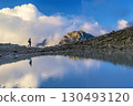 A person stands on rocky terrain, gazing at a stunning turquoise mountain lake surrounded. Dressed in hiking gear and holding trekking poles. Wildsee Pizol, Switzerland. 130493120