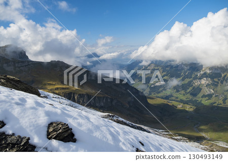 Snow blankets the rocky peaks of the Swiss Alps, contrasting with lush green valleys below. Fluffy clouds drift lazily across a stunning blue sky, creating a picturesque landscape. 130493149