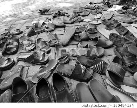 A chaotic jumble of various sandals and flip-flops, captured in a black and white image, scattered on a brick-paved ground. 130494184