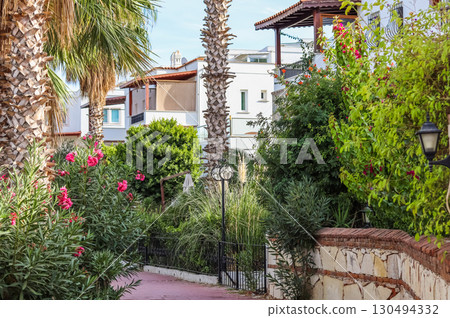 A narrow, winding path leads past lush green bushes and vibrant pink flowers, with white Mediterranean style homes and palm trees in the background. 130494332