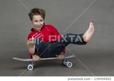 Funny little boy sitting on a vintage skateboard barefoot on a gray background. 130494892