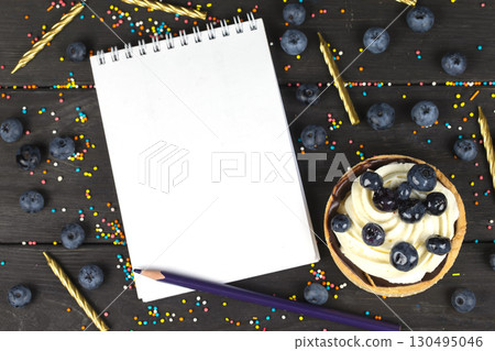 On a wooden board lies a cake with cream and blueberries and a white notebook with blank pages. On a wooden board lies a cake with cream and blueberries and a white notebook with blank pages. 130495046