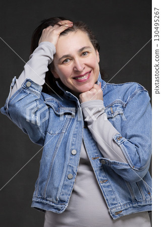 Happy fifty year old woman with dark hair in a denim jacket on a gray background. Portrait of a middle-aged woman. 130495267
