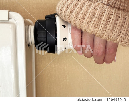 Woman hand in warm sweater unscrews radiator valve to turn on heating. Cold weather, beginning of heating season, regulating room temperature, warmth and comfort at home concept Woman hand in warm sweater unscrews radiator valve to turn on heating. Cold weather, beginning of heating season, regulating room temperature, warmth and comfort at home concept 130495943