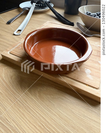 A round brown ceramic baking dish stands on a wooden cutting board in the kitchen. Cookware, home cooking, and culinary preparation reflect lifestyle, culture, and household traditions. 130496022