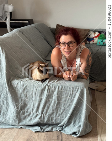A woman with glasses and tattoos lies on a sofa next to her siamese cat. Home atmosphere, companionship, and lifestyle moments reflect connection, relaxation, and daily human-animal bond. 130496023