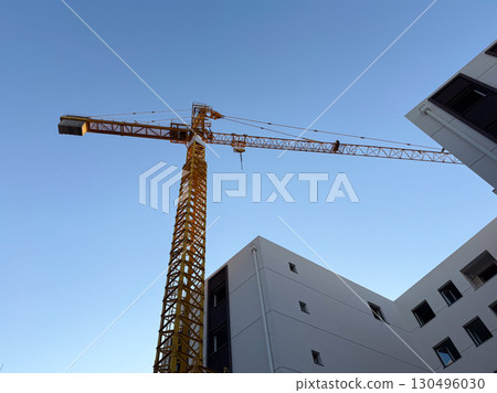 A tall construction crane rises above modern residential buildings against a clear sky. Engineering, infrastructure, and urban development highlight industrial growth and real estate progress. 130496030
