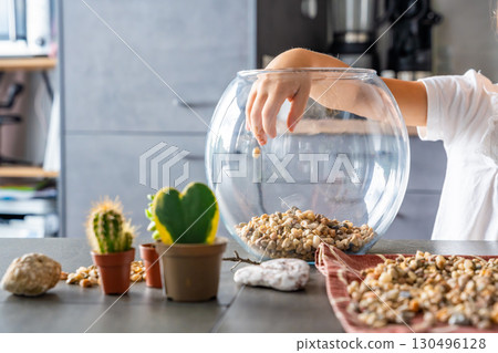 Close-up of child hands pouring stones into a glass container while preparing drainage for a florarium or closed ecosystem. Tactile learning, mindful creativity and eco-friendly crafting. 130496128