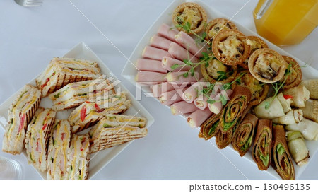 Gourmet catering spread featuring club sandwiches, ham rolls, savory pies, crepe wraps garnished with pea sprouts, plated on white rectangles near fresh juice pitcher Gourmet catering spread featuring club sandwiches, ham rolls, savory pies, crepe wraps garnished with pea sprouts, plated on white rectangles near fresh juice pitcher 130496135