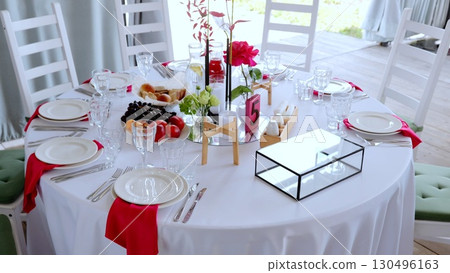 White draped round table featuring floral centerpiece, scattered fruits, numbered five, set for elegant outdoor dining event with sophisticated reception styling 130496163