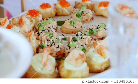 Elegant canapes arranged on a white plate, featuring toppings like shrimp, cucumber, sesame seeds, and creamy spread, perfect for festive gatherings 130496165