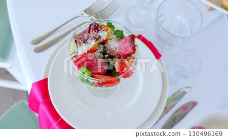 Glass bowl containing savory beef salad, sprinkled with parmesan, accompanied by ripe tomatoes and fresh arugula, resting on crisp white dinnerware with elegant table setting 130496169