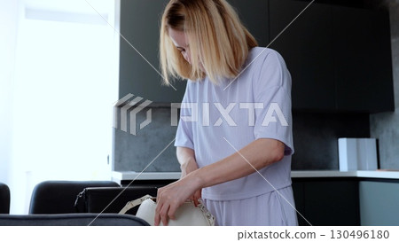 Young blond woman in a modern apartment, casually searching her handbag in the natural light before starting her day 130496180