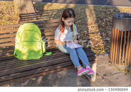 Young girl with backpack sitting on park bench, writing in notebook on autumn day 130496694