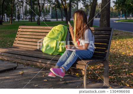 Girl with backpack relaxing on park bench using smartphone on a sunny day Girl with backpack relaxing on park bench using smartphone on a sunny day 130496698