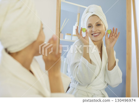 Smiling woman holding two cucumber slices near face, going to apply on eyes for skin moisturising 130496974
