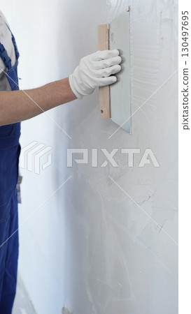 Male construction worker wearing protective gloves and blue construction coveralls, spreading plaster smoothly across wall using professional drywall taping knife during renovation, vertical view 130497695