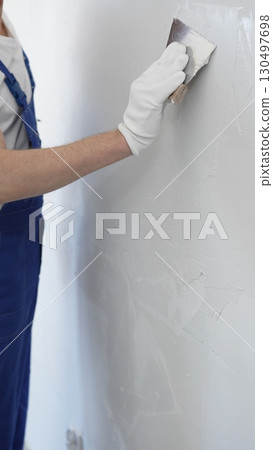 Close-up of man construction worker wearing gloves and blue overalls is using a putty knife to apply plaster on a white wall, performing home renovation and improvement Close-up of man construction worker wearing gloves and blue overalls is using a putty knife to apply plaster on a white wall, performing home renovation and improvement 130497698