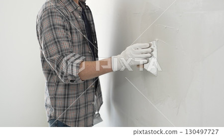 Unknown construction worker man wearing gloves and plaid shirt is using a putty knife to apply plaster on a white wall, during a home renovation or construction project 130497727