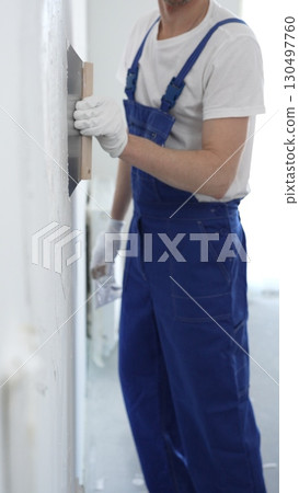 Man construction worker wearing protective gloves and blue construction coveralls, spreading plaster smoothly across wall using professional drywall taping knife 130497760
