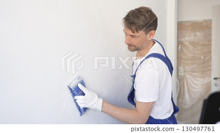 Man construction worker wearing protective white gloves and blue construction coveralls, is sanding white wall with blue hand block, preparing smooth surface during interior home renovation project 130497761