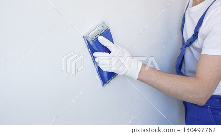 Unknown male construction worker wearing protective white gloves and blue construction coveralls, is sanding white wall with blue hand block, preparing smooth surface during interior home renovation 130497762