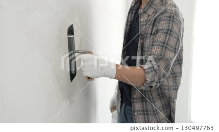 Unknown male construction worker wearing gloves and plaid shirt is using finishing trowel to plaster a white wall, applying a layer of plaster to create a smooth surface during renovation project 130497763