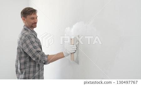 Man construction worker wearing protective gloves and plaid shirt, spreading plaster smoothly across wall surface using professional drywall taping knife during renovation project 130497767