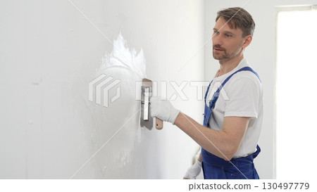 Professional male construction worker is using a finishing trowel to carefully apply plaster on a wall, demonstrating expertise in home renovation and interior finishing. Portrait view Professional male construction worker is using a finishing trowel to carefully apply plaster on a wall, demonstrating expertise in home renovation and interior finishing. Portrait view 130497779