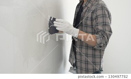 Unknown construction worker man wearing gloves and plaid shirt is using a putty knife to apply plaster on a white wall, during a home renovation or construction project 130497782