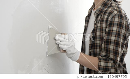 Unknown female construction worker wearing protective gloves and checkered shirt is spreading plaster on white wall during renovation project, smoothing surface with putty knife 130497795