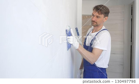 Man construction worker wearing protective white gloves and blue construction coveralls, is sanding white wall with blue hand block, preparing smooth surface during interior home renovation project 130497798