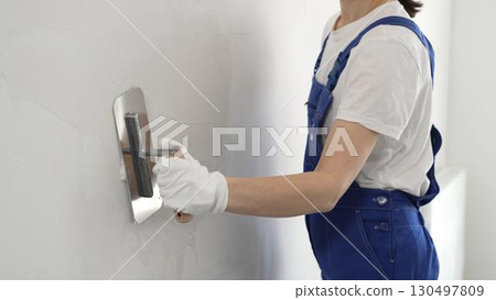 Professional female construction worker is using a finishing trowel to carefully apply plaster on a wall, demonstrating expertise in home renovation and interior finishing 130497809