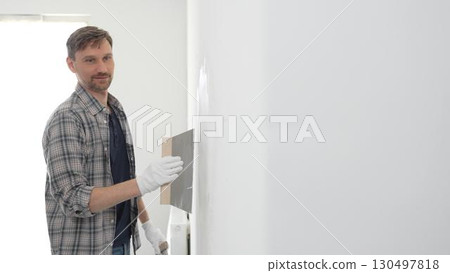 Man construction worker wearing protective gloves and plaid shirt, spreading plaster smoothly across wall surface using professional drywall taping knife during renovation project 130497818