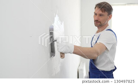 Professional male construction worker is using a finishing trowel to carefully apply plaster on a wall, demonstrating expertise in home renovation and interior finishing. Portrait view Professional male construction worker is using a finishing trowel to carefully apply plaster on a wall, demonstrating expertise in home renovation and interior finishing. Portrait view 130497825