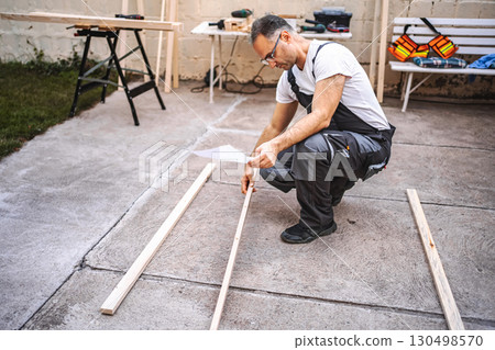 Mature male carpenter using electric screwdriver on a wooden plank outdoors 130498570
