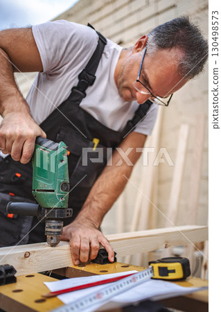 Mature male carpenter using electric screwdriver on a wooden plank outdoors Mature male carpenter using electric screwdriver on a wooden plank outdoors 130498573